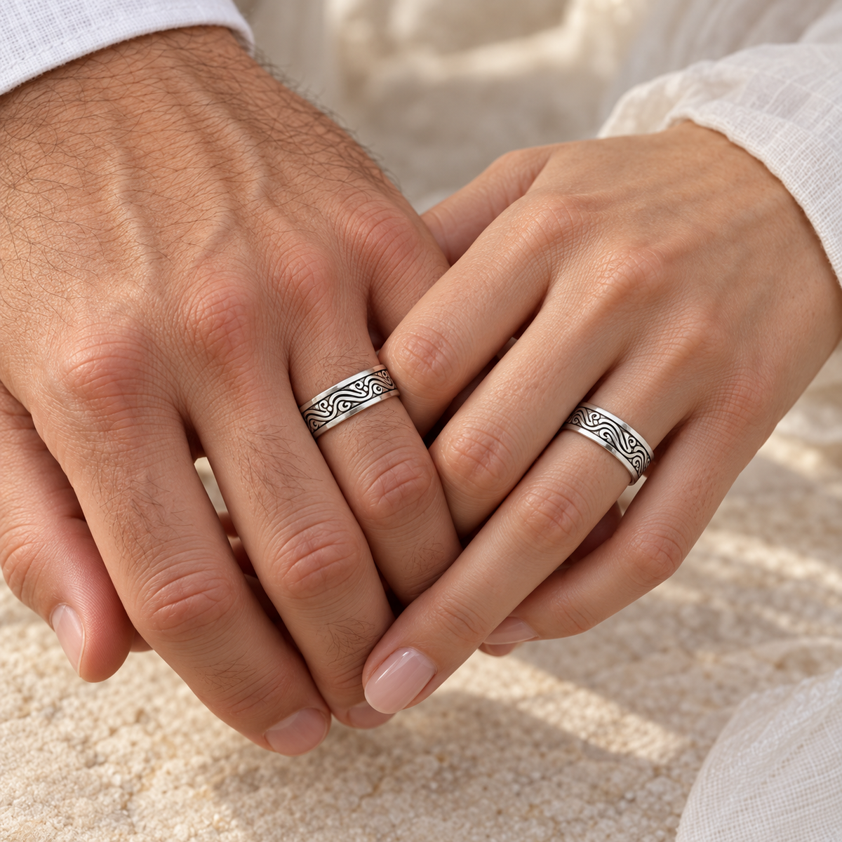 Two hands clasped together wearing matching platinum rings with an engraved wavy pattern, set against a soft beige background.