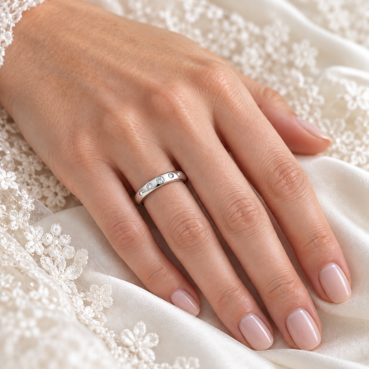 A close-up of a hand wearing a slim platinum wedding band ring set with diamonds, resting on white lace fabric.